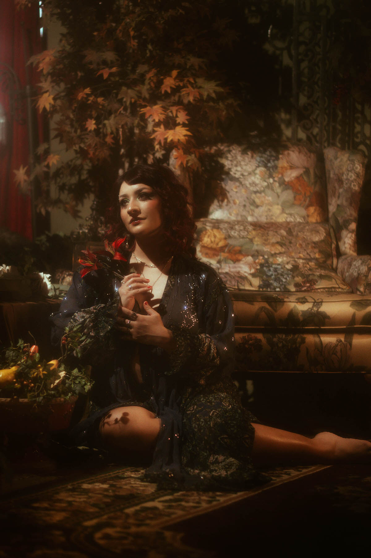 Whimsical maternity portrait of a muse in a sparkling navy robe, seated on an antique floral couch, gently holding a red flower with soft, romantic lighting at Royal Lune Studio.