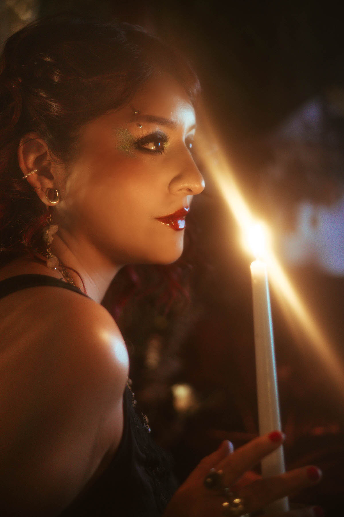 A close-up side profile of a woman with red and black curled hair, holding a single white taper candle in a brass holder. The candle casts a warm glow on her face, highlighting her shimmering green eye makeup and deep red lips. The scene is set against a dark, ethereal backdrop with blurred hints of floral elements and antique decor, evoking a sense of mystery and elegance.