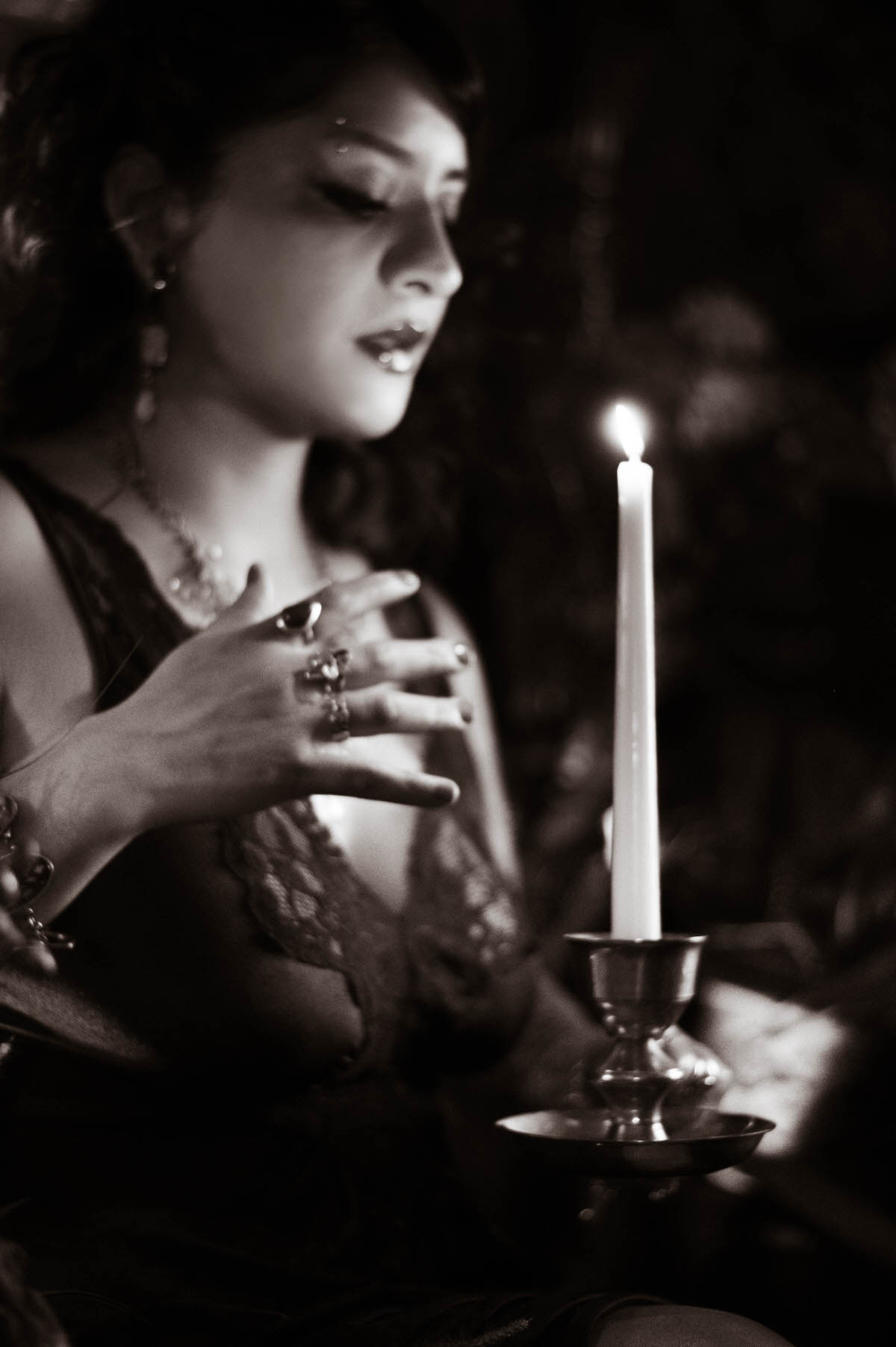 A dramatic black-and-white image of a pregnant woman in a lace dress, gently touching her candlelit face with one hand. The scene captures the play of shadows and highlights, emphasizing the delicate textures of the lace and the soft curve of her baby bump. The timeless, moody tone adds a sense of quiet introspection.