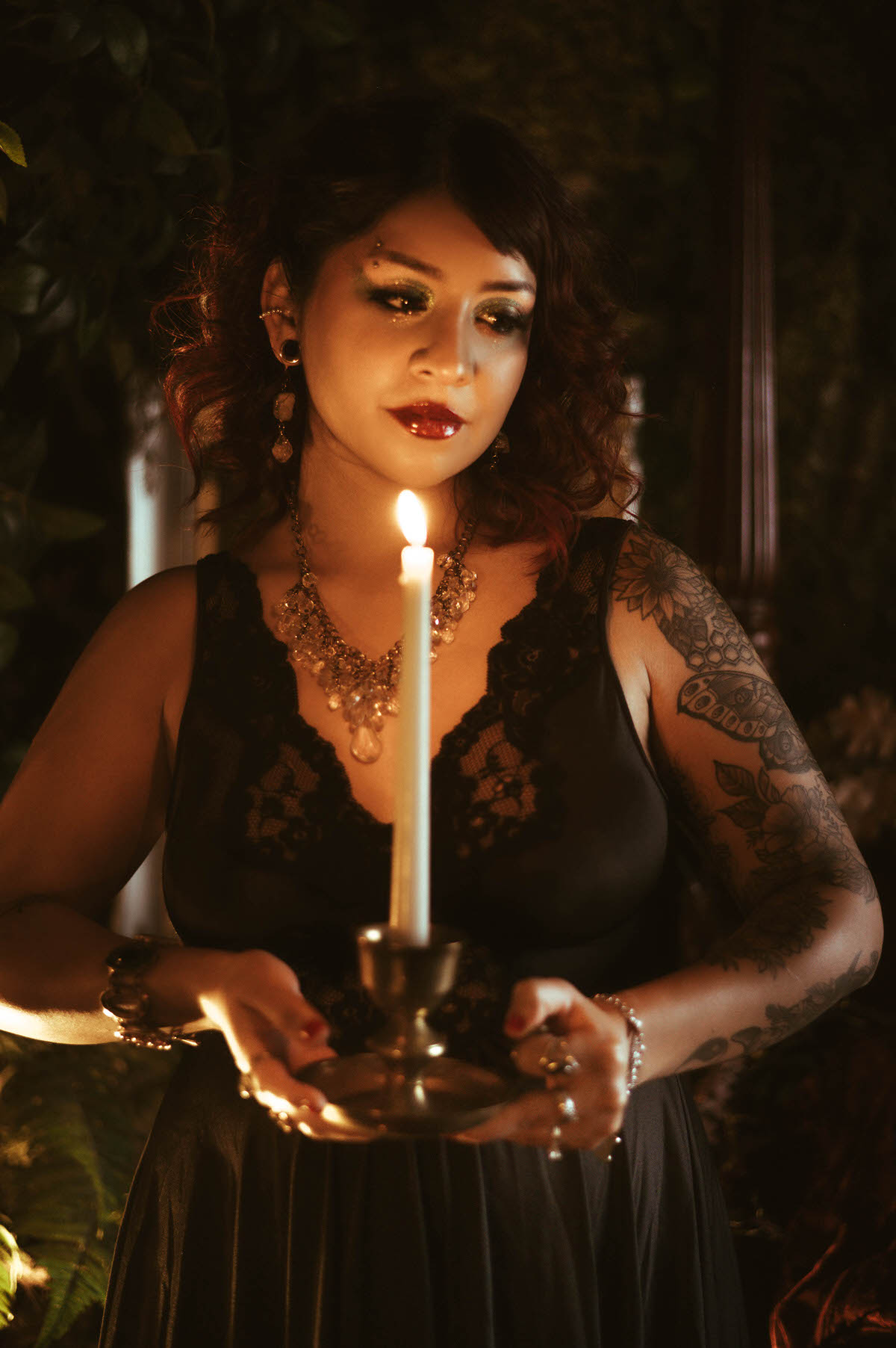 A maternity portrait featuring a woman in a black lace vintage nightgown, standing in a lush, enchanted garden setup. She holds a white taper candle in a brass holder, her gaze downcast as the warm candlelight illuminates her face and ornate crystal necklace. The scene is filled with rich shadows, deep greenery, and romantic, antique-inspired details.