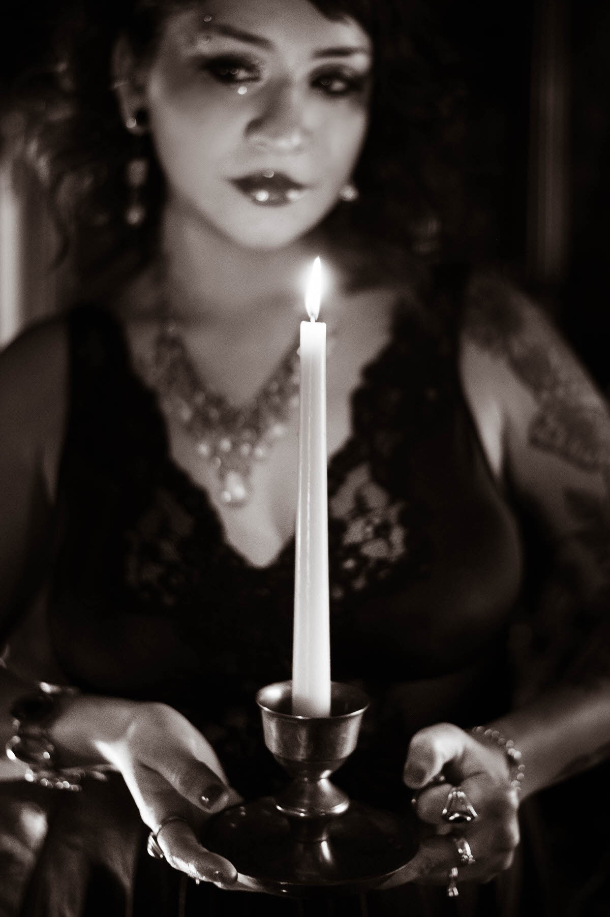 An evocative portrait of a woman in a black lace nightgown, holding a white taper candle as she stands in front of a lush, green backdrop. The candlelight highlights her vibrant red curls and glittering crystal necklace, casting a warm, golden glow that contrasts with the deep, rich shadows of the scene.