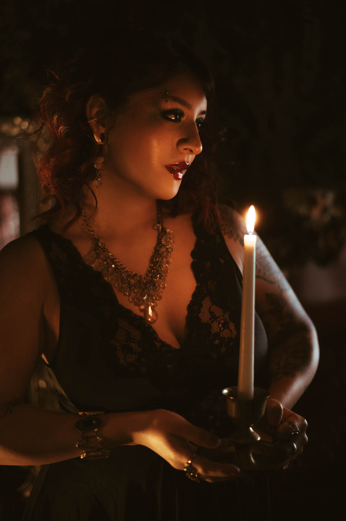 A close-up portrait of a woman with curled red and black hair, illuminated by the golden glow of a candle. Her eyes are focused on the flame, creating a contemplative, almost mystical atmosphere. The background is filled with dark greenery and soft, defocused lights, enhancing the sense of intimacy and wonder.
