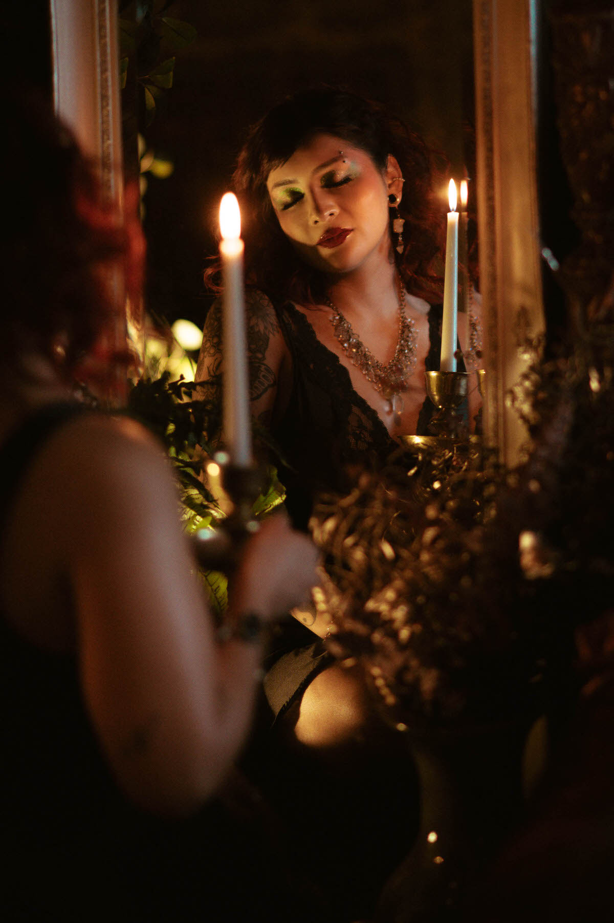 An artistic reflection shot, capturing a woman holding a brass candle holder while gazing into a vintage mirror. The warm candlelight casts a soft, flattering glow on her face, emphasizing her dramatic green eyeshadow and crimson lips. The surrounding floral arrangements and antique frame add layers of depth and texture to the scene.