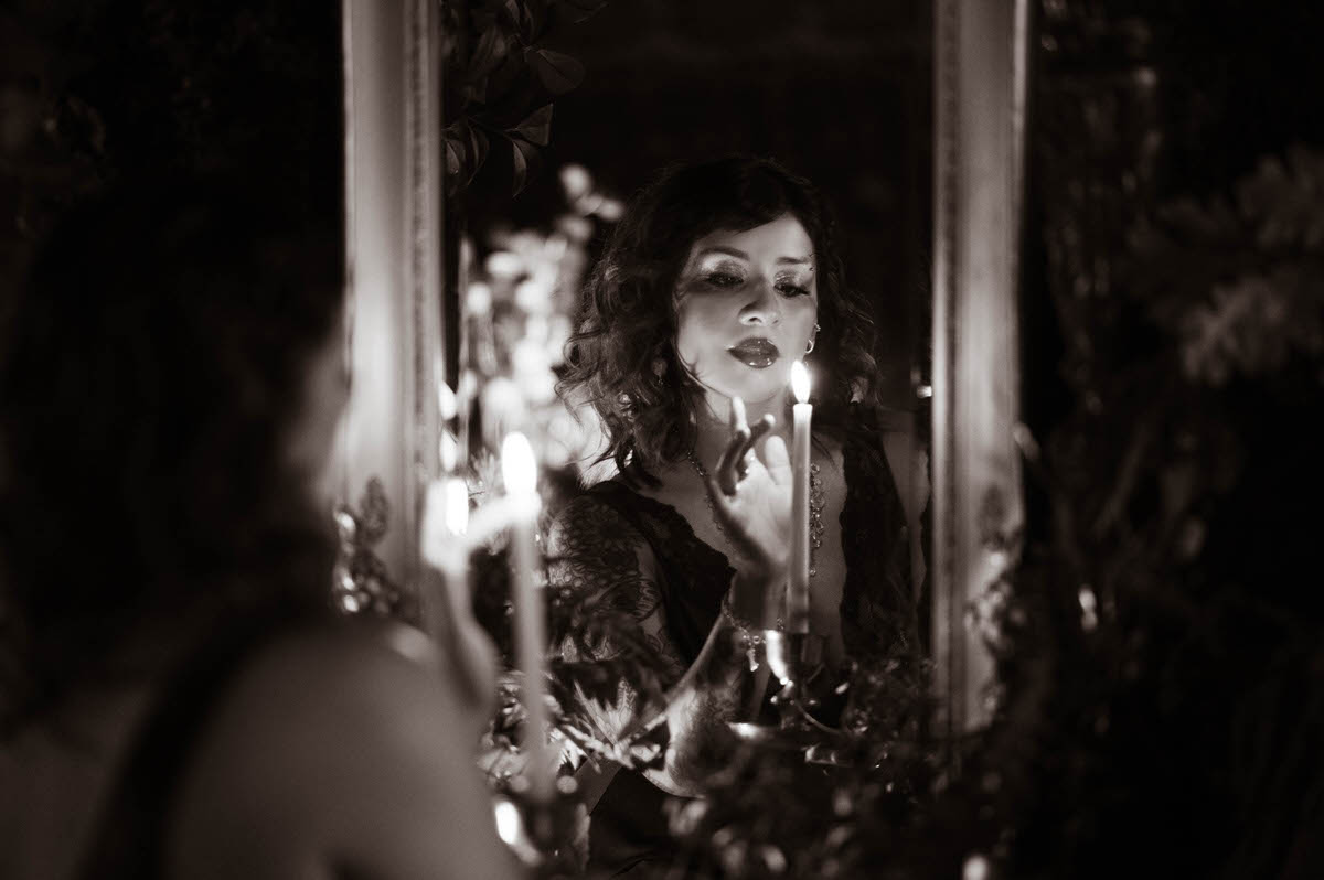 A moody, ethereal portrait of a woman in a black lace nightgown, holding a tall white taper candle with a brass holder. The soft, warm candlelight illuminates her face, casting delicate highlights on her skin and creating dramatic shadows against the lush, botanical backdrop, evoking a mystical, timeless atmosphere.