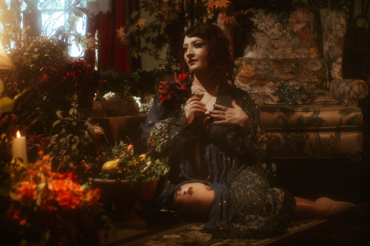 Ethereal maternity boudoir portrait of a muse resting against a floral tapestry backdrop, surrounded by lush florals and candlelight, wearing a sheer, sequin-studded navy robe at Royal Lune Studio.