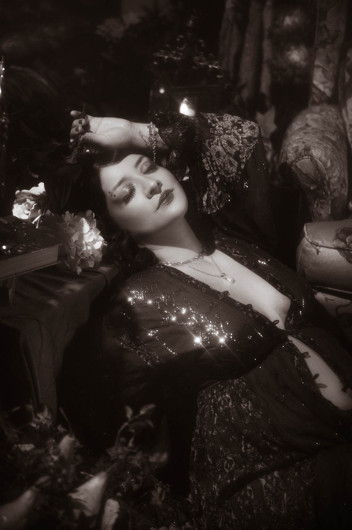 Vintage-inspired maternity portrait in black and white, featuring a muse in a flowing, sheer gown, resting her head against a pile of antique books, surrounded by romantic florals for a moody, cinematic maternity look. 