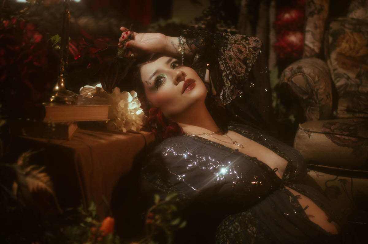 Dreamy maternity scene featuring the muse reclining against a vintage tapestry-covered sofa, draped in a shimmering blue gown, with flickering candlelight and textured florals enhancing the moody, cinematic atmosphere at Royal Lune Studio.