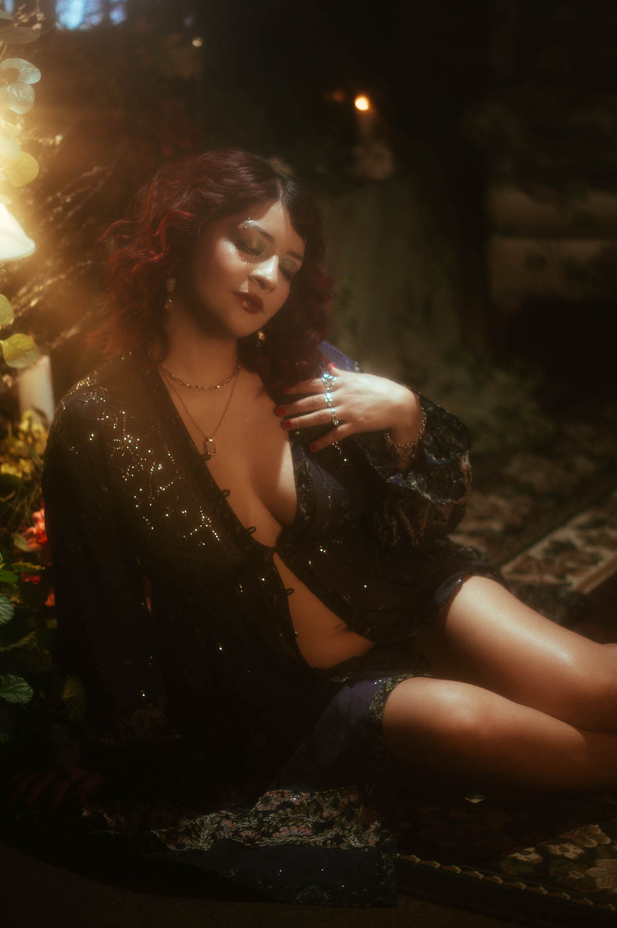 A moody maternity portrait with romantic styling, capturing the mother-to-be draped in a sheer, sequin-embellished gown, her hand gently resting on her growing belly, surrounded by lush florals and soft candlelight at Royal Lune Studio.