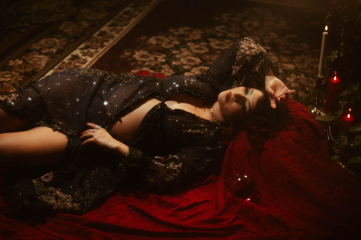 A dreamy, vintage-inspired maternity portrait featuring a woman with soft red curls, glowing green eyeshadow, and a shimmering gold dress. She stands beside a tall, ornate column with a classical Grecian vase, surrounded by lush greenery and warm, romantic candlelight, capturing the ethereal essence of fine art photography.