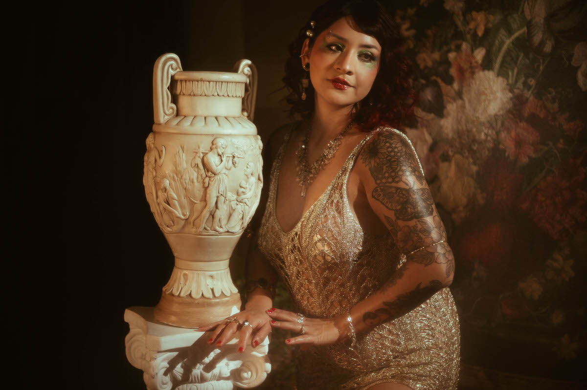 A close-up portrait of a woman with vibrant red curls and striking green eyeshadow, her eyes gently closed as she leans against a Grecian vase. Her shimmering gold dress and intricate jewelry catch the warm, romantic light, highlighting the fine art details of this intimate maternity scene.