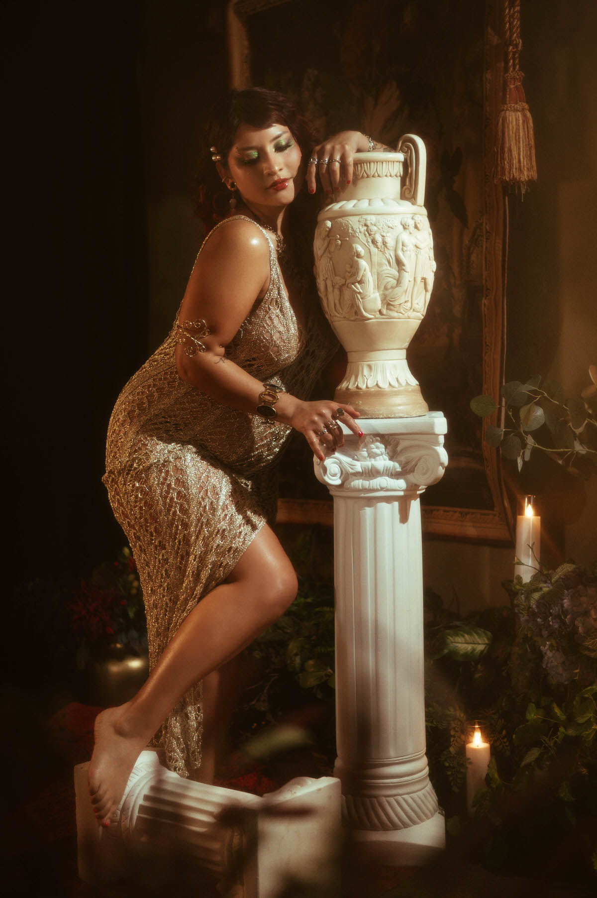 A dreamy, vintage-inspired maternity portrait featuring a woman with soft red curls, glowing green eyeshadow, and a shimmering gold dress. She stands beside a tall, ornate column with a classical Grecian vase, surrounded by lush greenery and warm, romantic candlelight, capturing the ethereal essence of fine art photography.