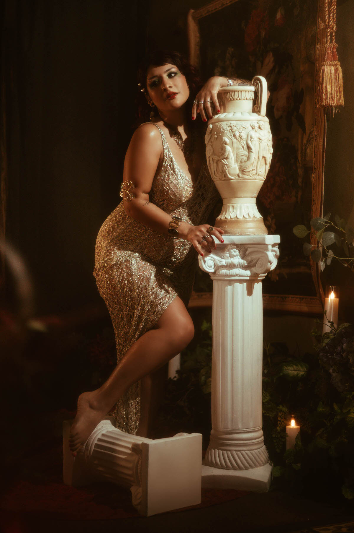 Maternity muse in a shimmering, gold crochet dress, leaning gracefully against a classical Greek column with ornate carvings, capturing the timeless beauty of artistic maternity portraits in a romantic, moody setting at Royal Lune Studio.