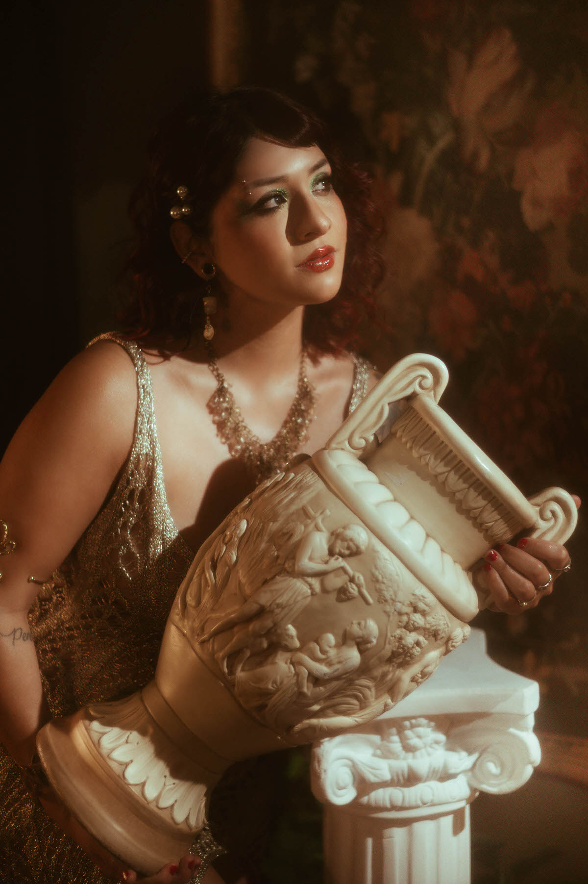 An intimate maternity portrait in a dark, romantic setting, featuring a woman with red curls and shimmering green eye makeup, holding a Grecian urn. The scene is bathed in soft, cinematic lighting, with rich textures and classical decor that evoke a sense of timeless elegance.