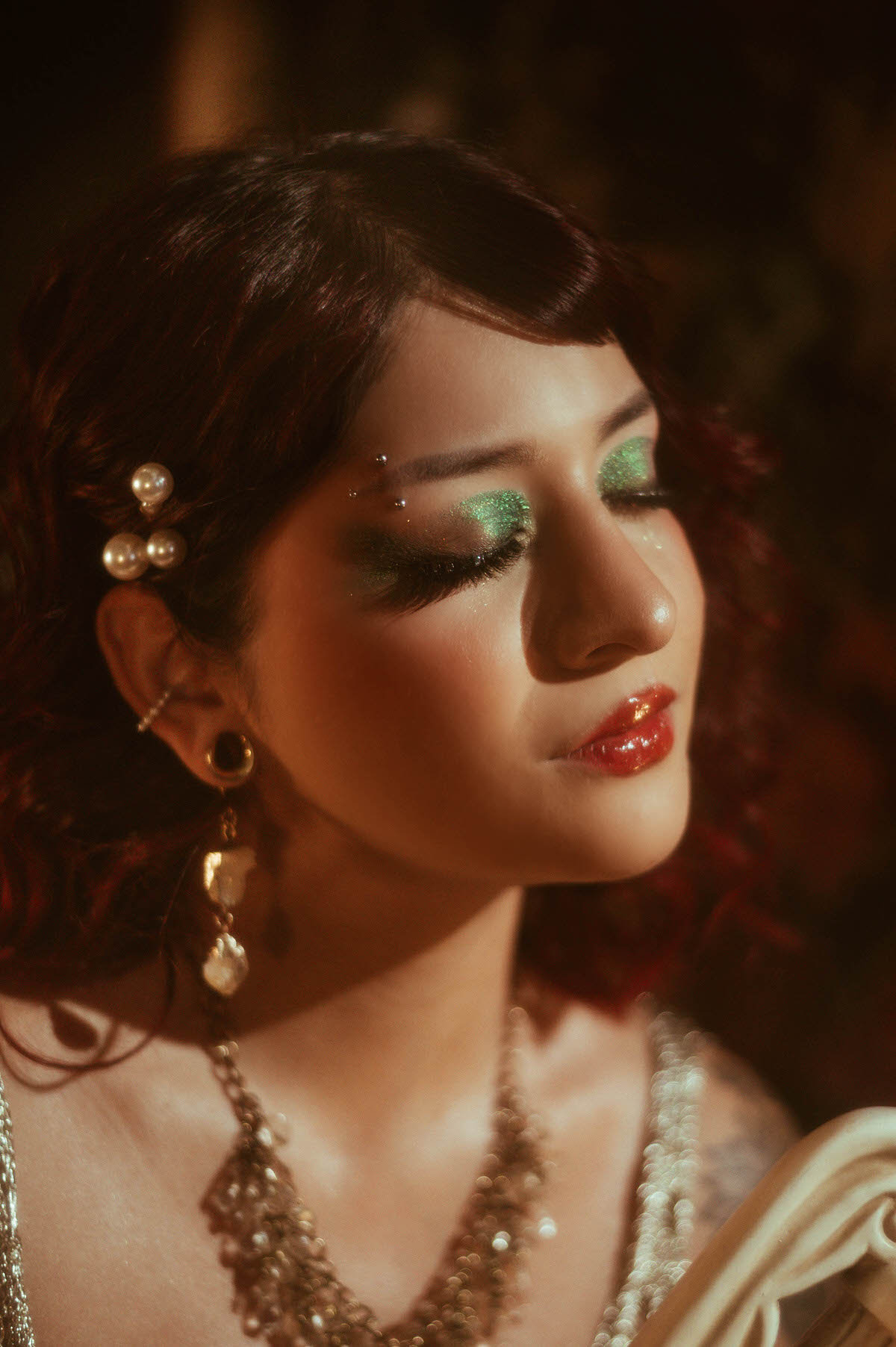 A golden hour portrait capturing the side profile of a woman with deep red curls, her eyes closed in a moment of quiet reflection. She wears a glittering gold dress and statement pearl accessories, standing beside a classical column with a detailed Grecian vase, surrounded by lush greenery and rich, warm tones.