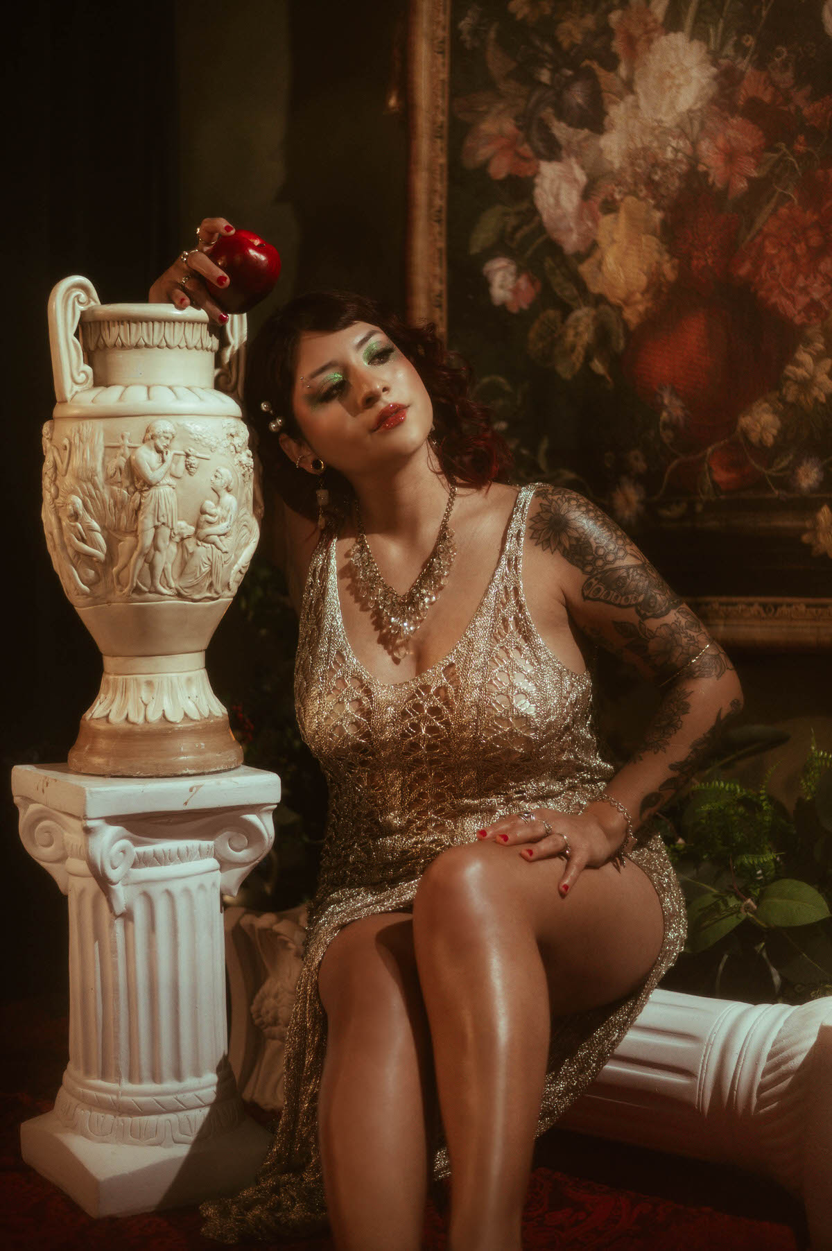 A pregnant woman in a shimmering, gold crochet dress sits gracefully on a vintage chaise lounge. She rests her hands on her belly, gazing softly to the side as warm golden light spills across her form, highlighting her intricate arm tattoos and layered jewelry.