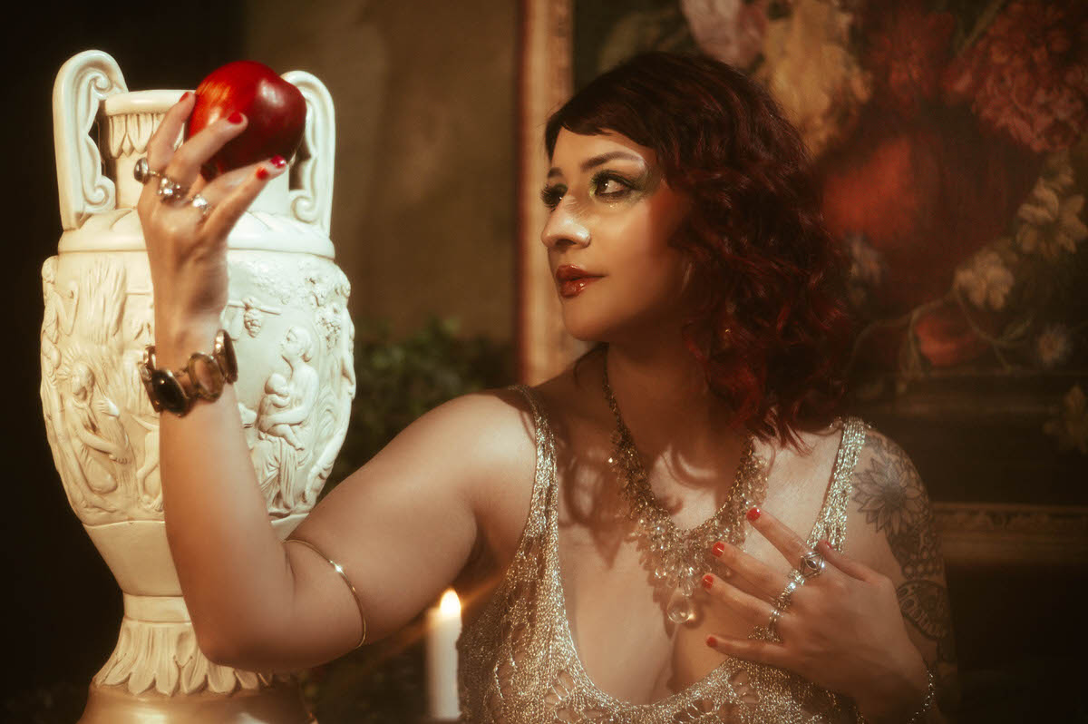 A close-up portrait of the woman with her eyes downward, her face illuminated by a warm, golden glow. Her richly textured dress and bold, glittering eye makeup add to the ethereal, cinematic feel of the image.