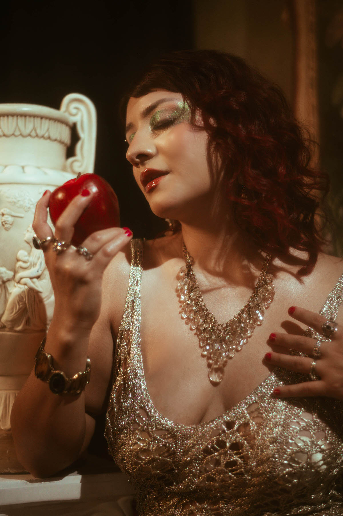 A close-up of the woman's face with her eyes closed, her dark lashes framed by vivid green eyeshadow. The lighting casts a warm glow across her serene expression, capturing the rich details of her intricate jewelry and softly curled red hair.