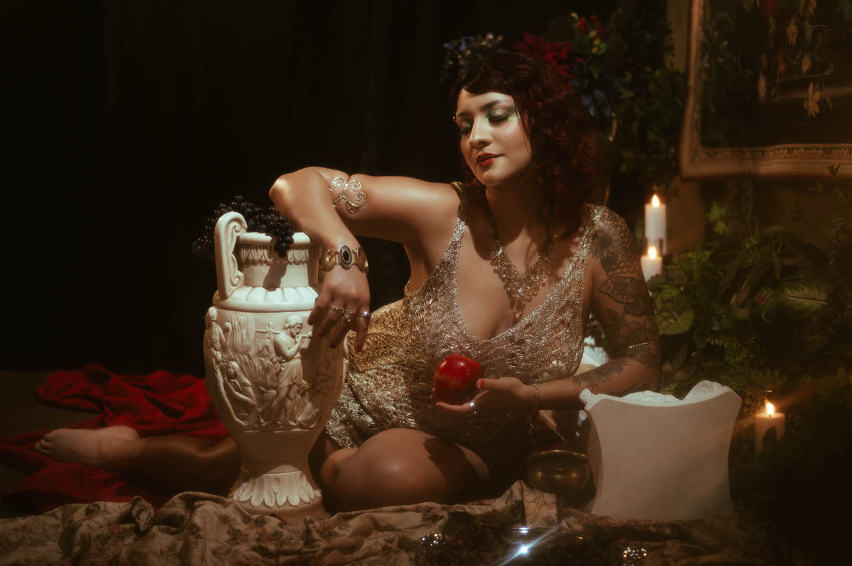 The woman reclines on a bed of deep red fabric, surrounded by lush greenery and ornate urns. Her gold crochet dress shimmers in the dramatic, low-key lighting, evoking a timeless, classical portrait style.