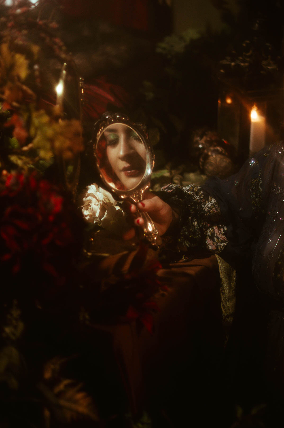 Dreamy maternity boudoir image of a woman gazing into an ornate vintage hand mirror, surrounded by red and white florals, soft candlelight, and romantic decor at Royal Lune Studio.