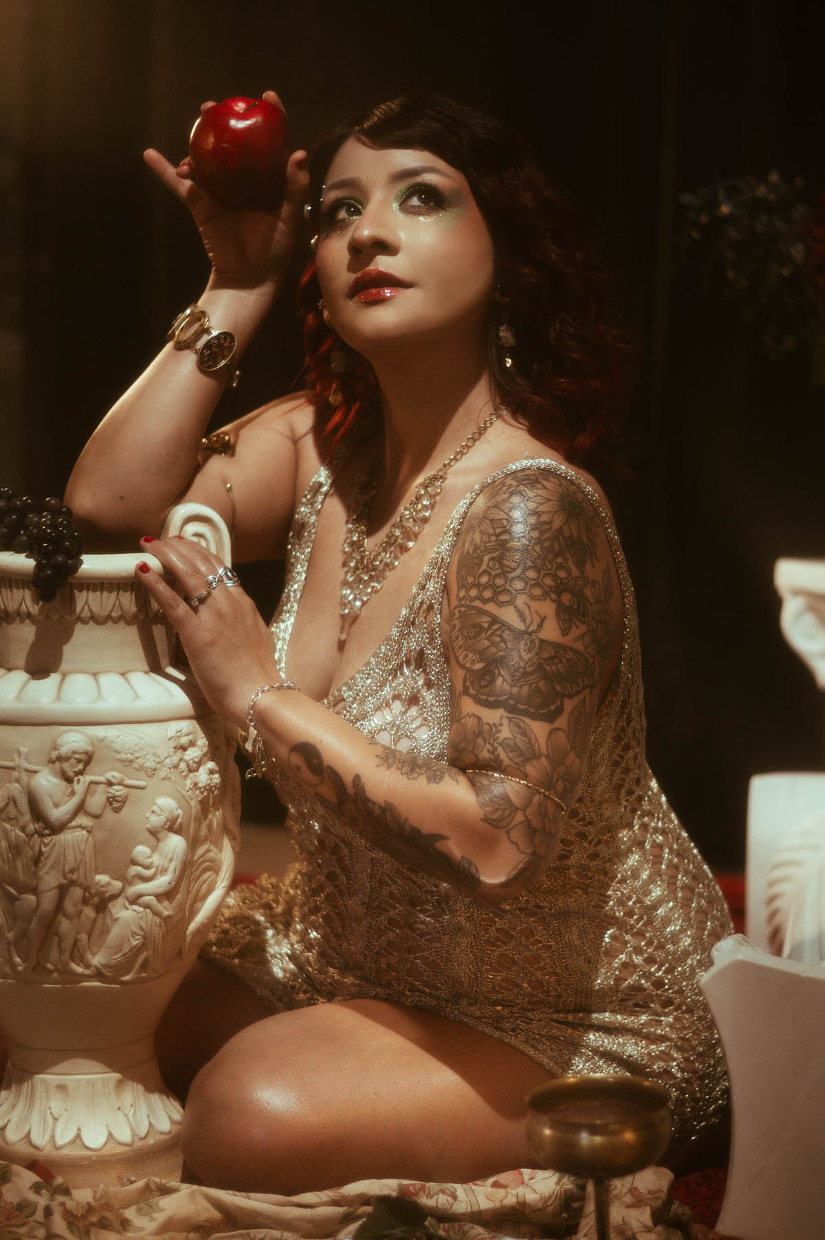 A pregnant woman in a shimmering, gold crochet dress sits gracefully on a vintage chaise lounge. She rests her hands on her belly, gazing softly to the side as warm golden light spills across her form, highlighting her intricate arm tattoos and layered jewelry.