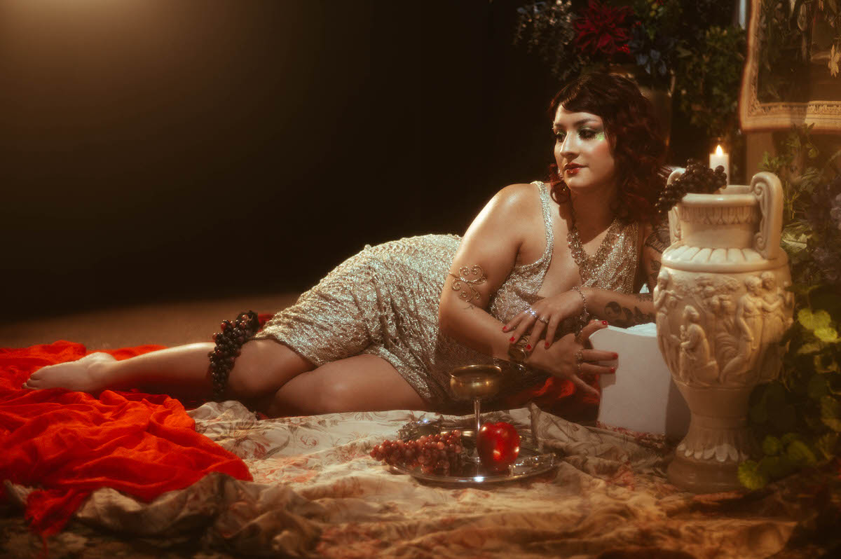 The woman lies on a deep, red velvet backdrop, her gold gown pooling around her as she reaches gracefully overhead, evoking the soft, flowing lines of Renaissance painting.
