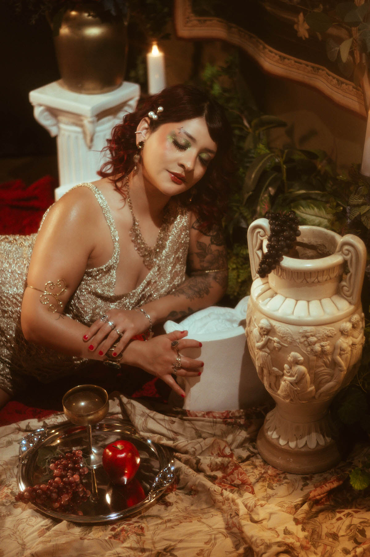 The woman kneels beside a white neoclassical urn, her gold gown catching the soft, cinematic light. Grapes spill over the urn, adding a touch of still life drama to the scene as she gazes thoughtfully at the fruit.