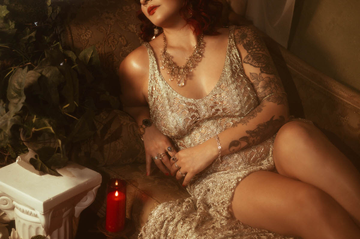 A pregnant woman in a shimmering, gold crochet dress sits gracefully on a vintage chaise lounge. She rests her hands on her belly, gazing softly to the side as warm golden light spills across her form, highlighting her intricate arm tattoos and layered jewelry.