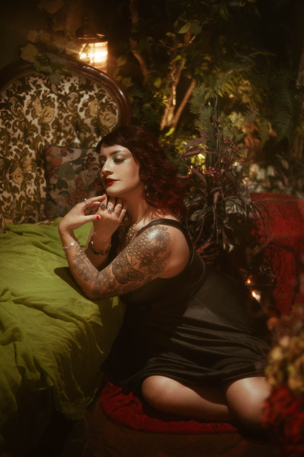 A dramatic maternity portrait with the muse seated on a vintage floral armchair, surrounded by lush greenery and soft, warm light. She wears a sheer black lace nightgown that cascades over the edge of the chair, her hands gently cradling her belly, capturing the essence of expectant motherhood in an ethereal, moody atmosphere.