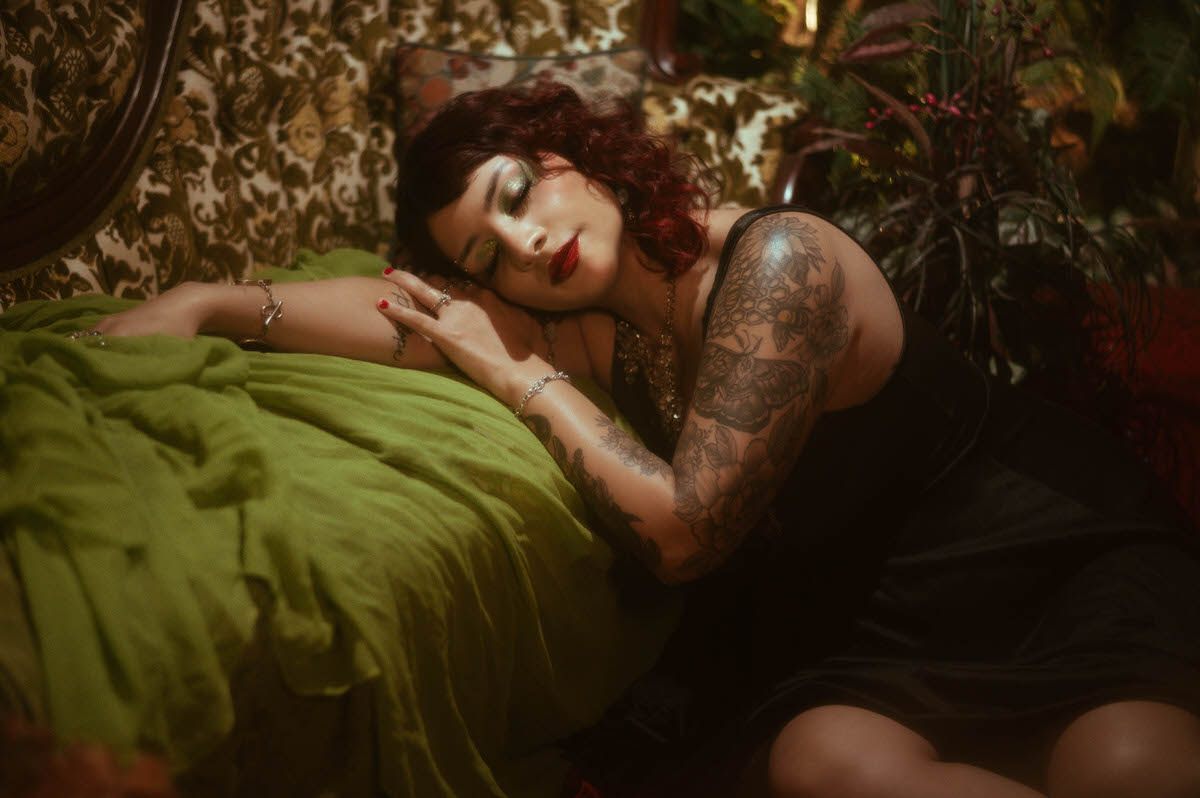 The muse leans forward in a thoughtful pose, her tattooed arms resting on a lush green velvet blanket draped over an antique armchair. Her gaze is lowered, eyes softly closed, with rich, moody lighting casting delicate shadows across her face, creating an intimate, reflective moment.