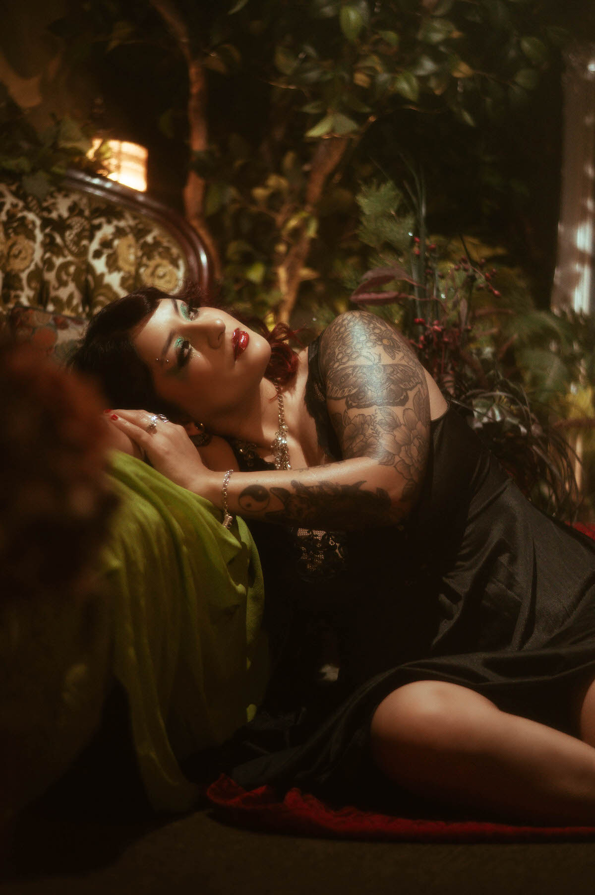 The muse reclines gracefully on an ornate vintage couch, surrounded by deep green foliage and atmospheric candlelight. She wears a black lace nightgown that softly clings to her form, with a calm, serene expression as she cradles her pregnant belly, capturing the essence of strength and maternal beauty.