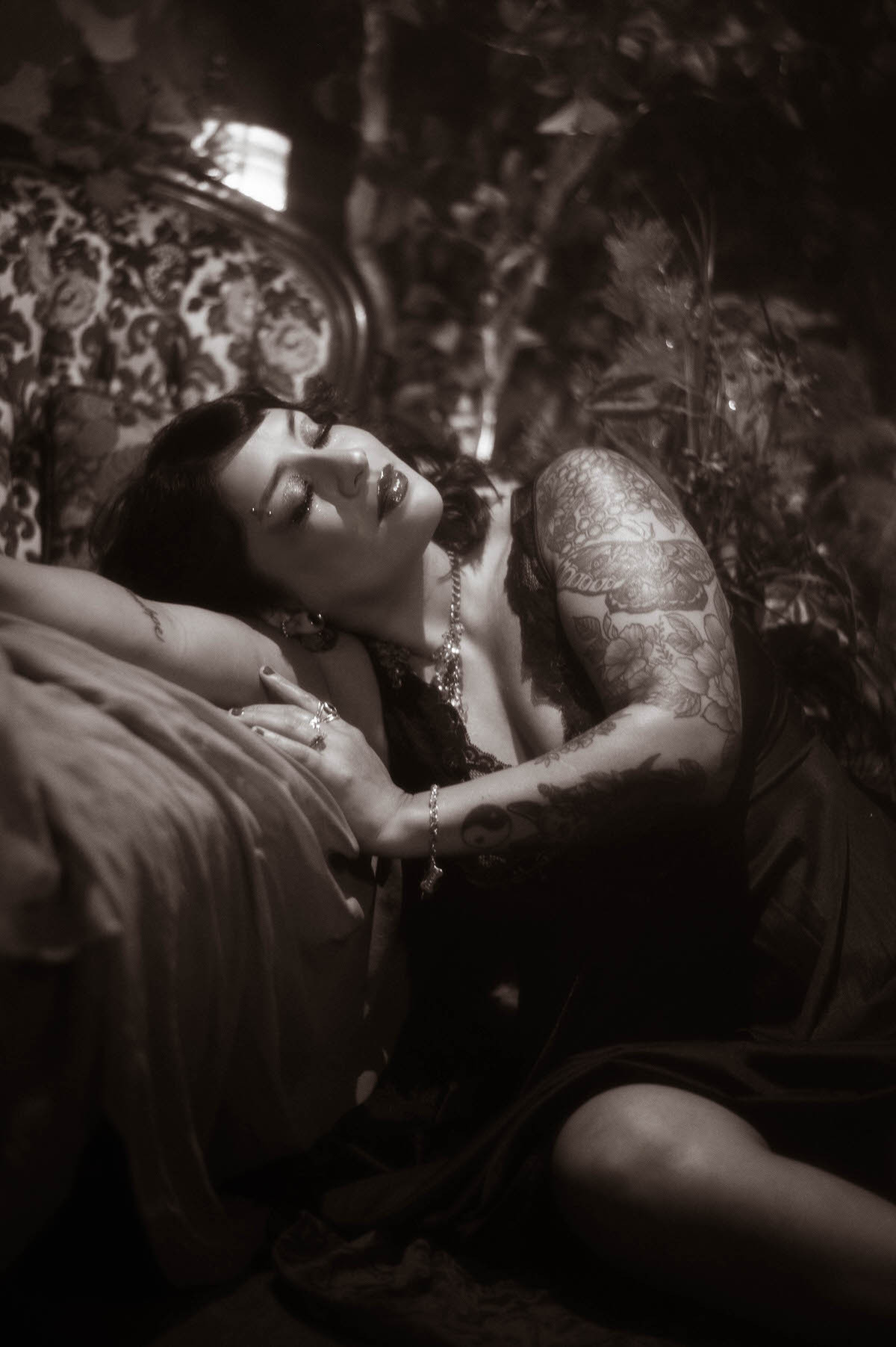 A soft, romantic black and white portrait of the muse leaning back on a vintage chair, eyes closed in a peaceful moment. Her dark hair cascades around her shoulders, and her lace gown adds a touch of gothic elegance to the scene, framed by moody, textured shadows.