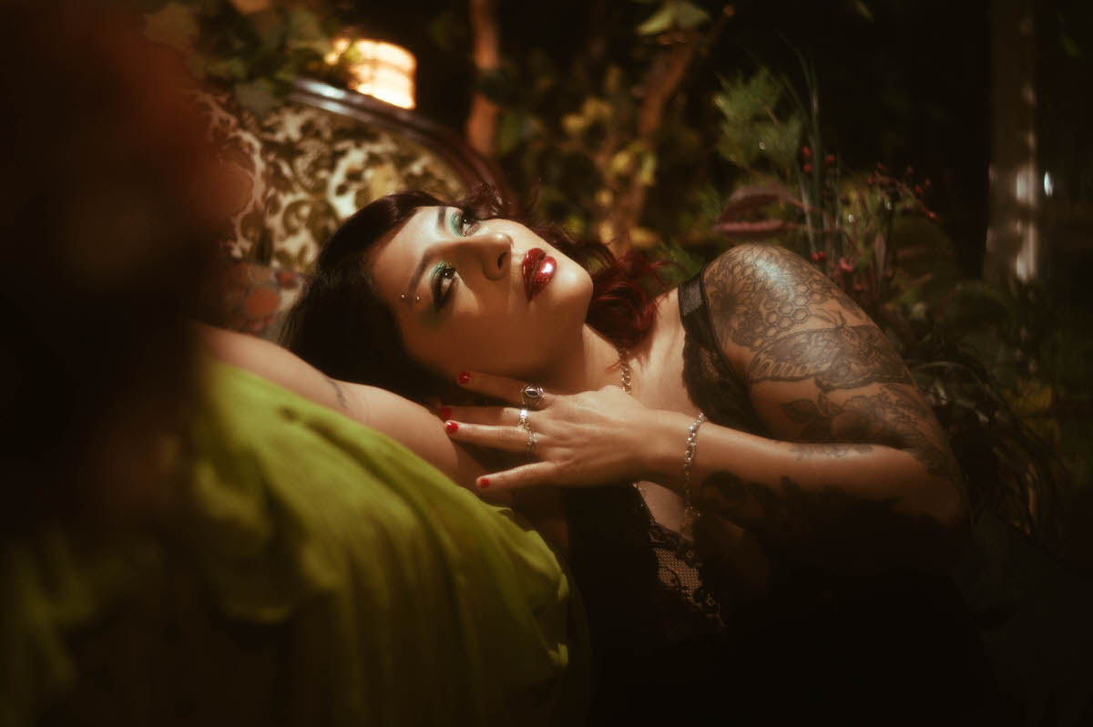 The muse rests against the lush greenery, her tattooed arm prominently displayed, adding a touch of artistic edge to the ethereal maternity scene. The warm, golden light highlights her serene expression, capturing the quiet strength and beauty of motherhood.