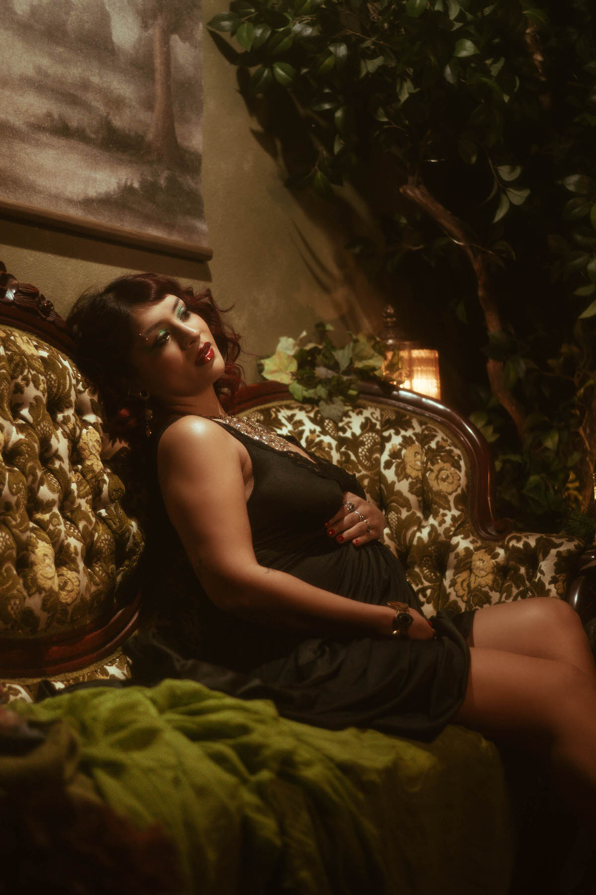 The muse lounges in a vintage-inspired setting, her head tilted back as she softly touches her belly, enveloped by cascading fabric and lush, ambient greenery. The scene is illuminated by the warm glow of a vintage lamp, enhancing the moody, intimate feel of the portrait.