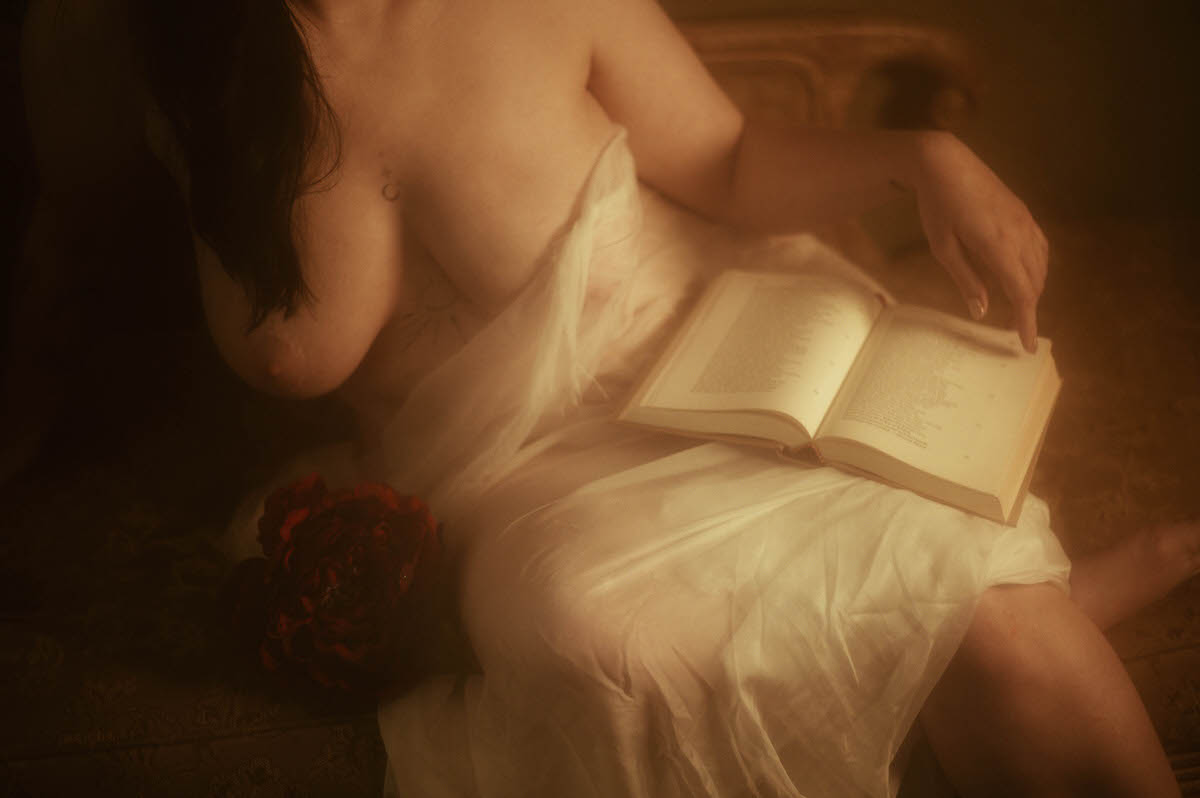 Painterly boudoir portrait of a partially nude woman draped in white fabric, seated on a vintage couch with an open book and red peony, capturing a soft, romantic moment of feminine stillness and intimacy.