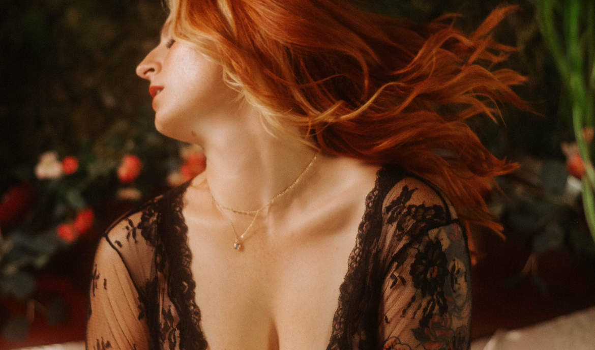 A red-haired woman in a black lace robe tosses her head gracefully, her hair caught mid-motion in a dreamy boudoir portrait. The romantic floral backdrop and soft lighting highlight her movement and neckline, illustrating dynamic boudoir posing for a fine art photography session.