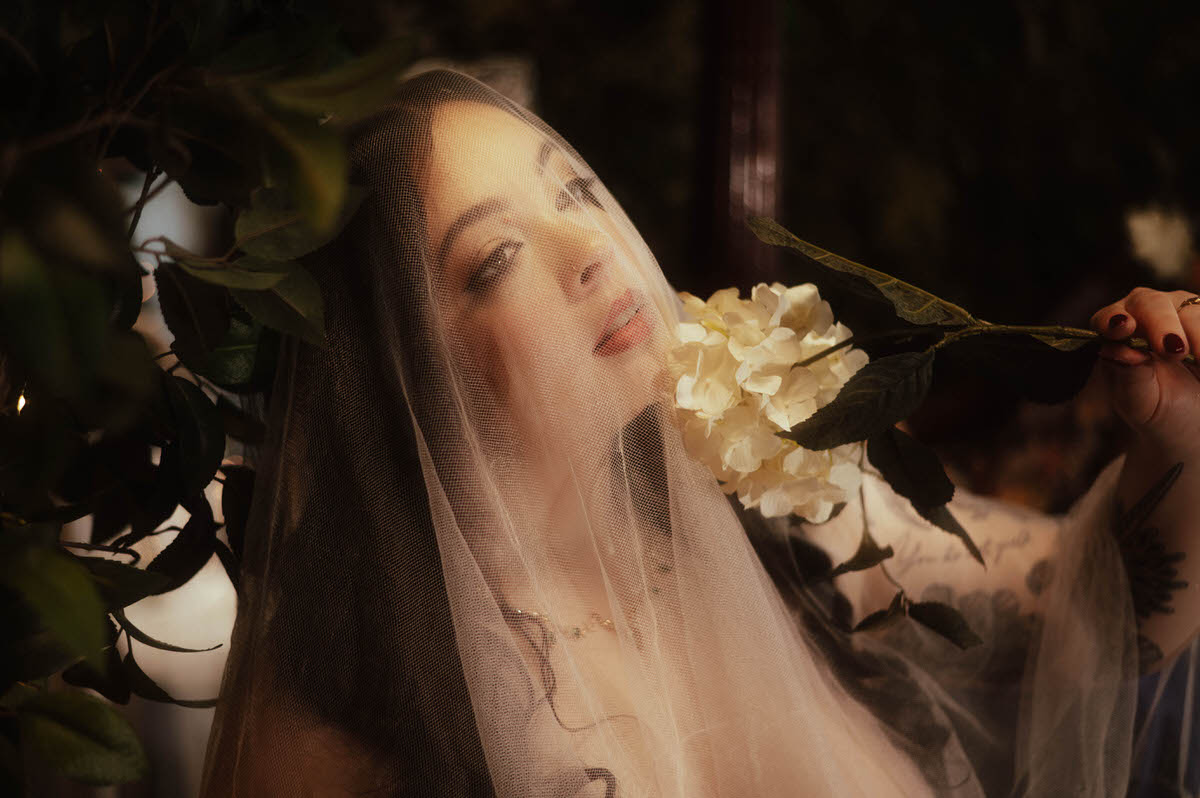 A dreamy boudoir portrait of a veiled woman softly gazing upward as she holds a white hydrangea bloom beside her face. Surrounded by greenery and candlelight at Royal Lune Studio, this image evokes moody boudoir photography with romantic, creative boudoir inspiration.