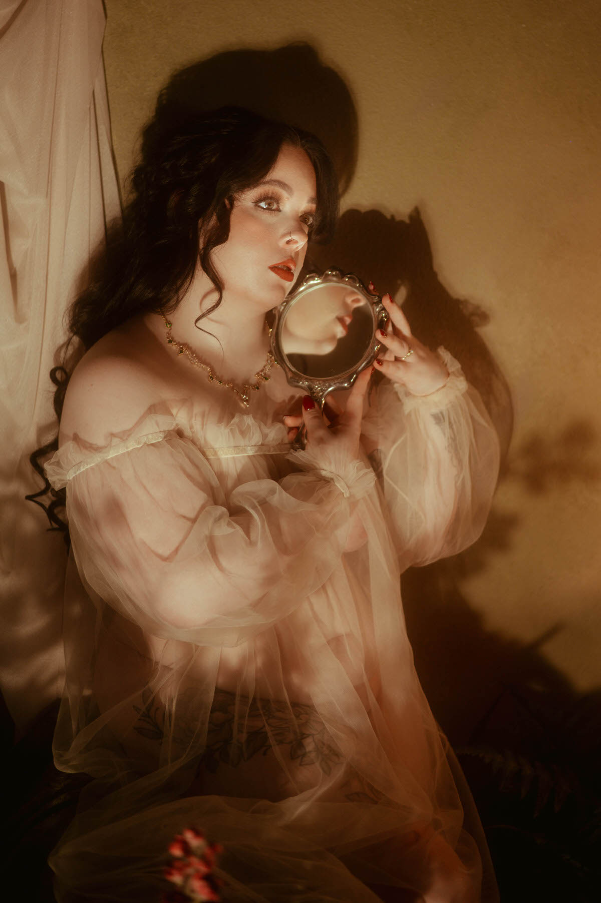A woman in a sheer off-the-shoulder tulle gown gazes into an ornate vintage hand mirror, her reflection partially visible as golden light and shadows play across her face and the wall. Captured at Royal Lune Studio, this romantic boudoir portrait evokes dreamy, fine art softness with vintage-inspired styling and ethereal mood.