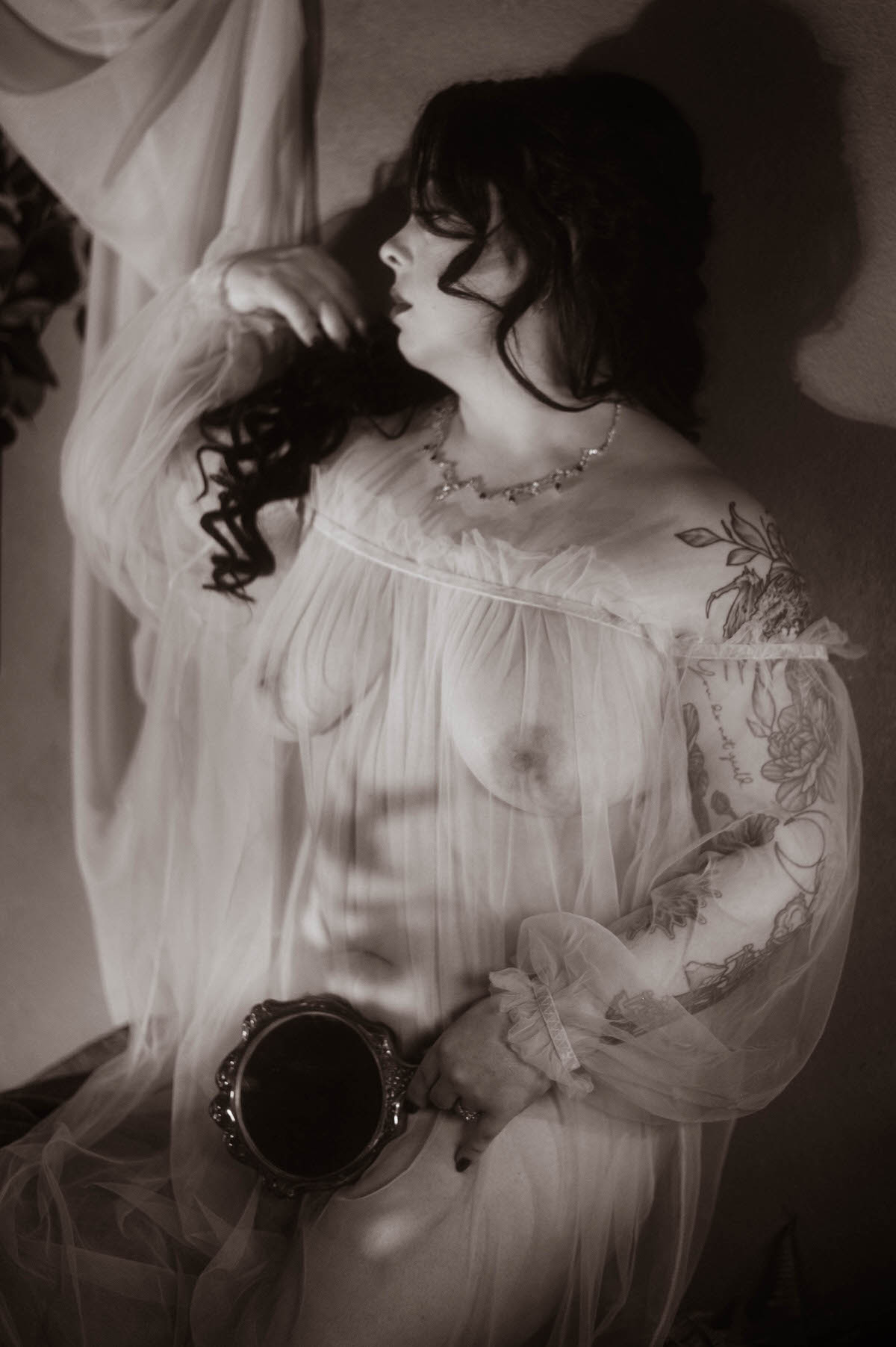 Black and white boudoir portrait of a woman in a sheer, off-the-shoulder gown, softly lit to reveal delicate shadows on her skin. She gazes to the side, one hand in her dark curled hair, the other resting on her hip while holding a vintage mirror. Her tattoos and translucent fabric evoke a romantic, ethereal aesthetic within the dreamy Royal Lune Studio setting.