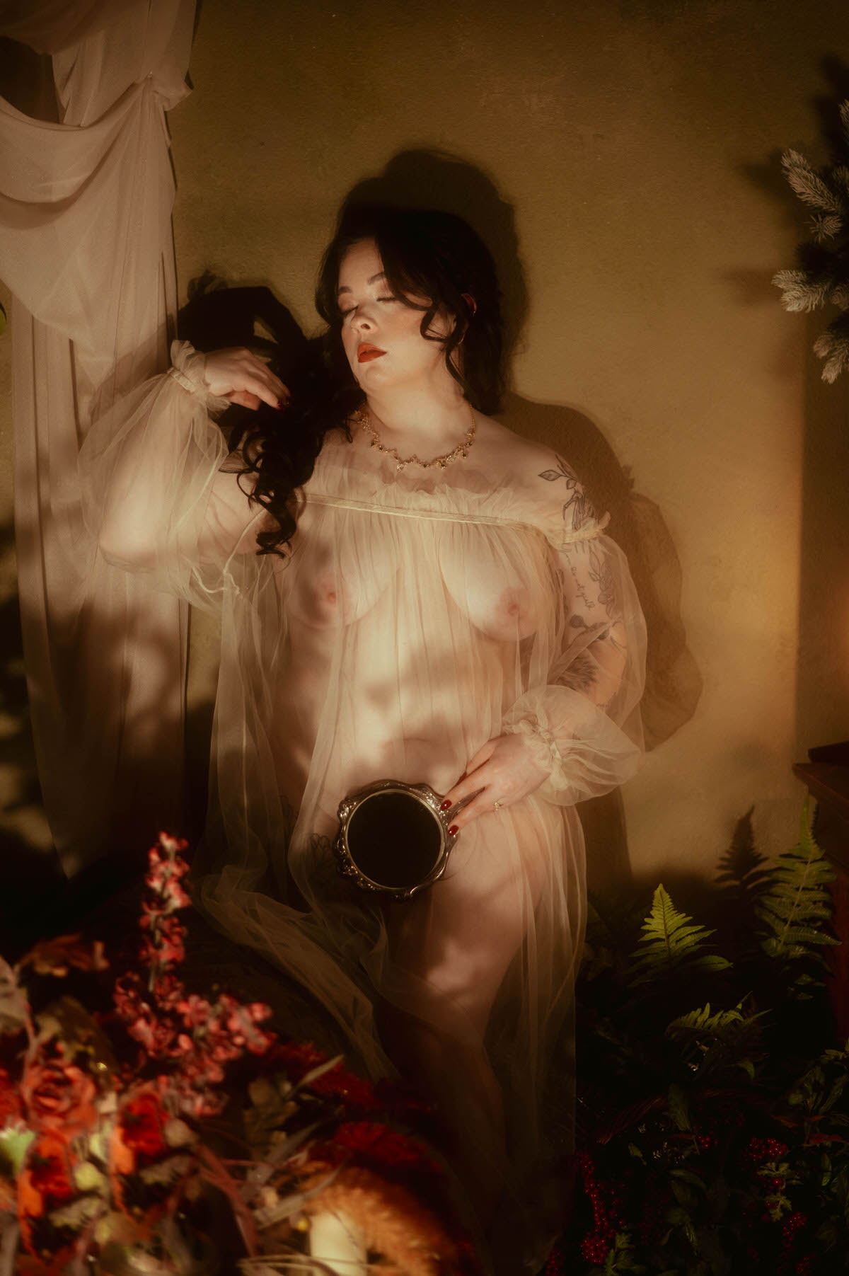 A woman stands in dreamy golden light wearing a sheer off-the-shoulder gown, holding a vintage mirror at her waist with one hand as the other brushes through her dark curls. The soft fabric reveals her form beneath, surrounded by rich florals and ferns at Royal Lune Studio. This fine art boudoir portrait channels romanticism and vintage elegance.