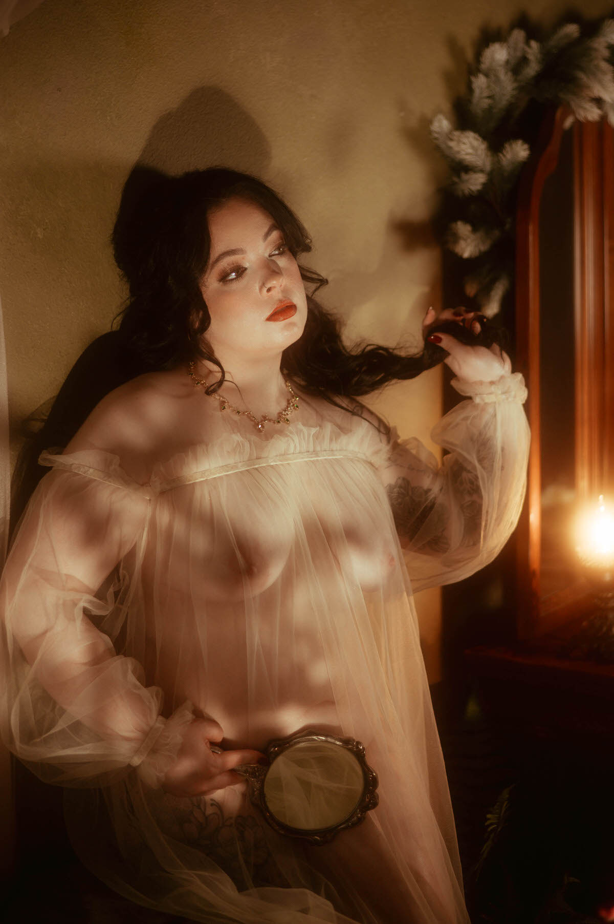 Dreamy boudoir portrait of a woman in a sheer, vintage-inspired gown, softly lit by candlelight at Royal Lune Studio. She holds a delicate hand mirror at her hip and gently twirls a lock of hair, her gaze cast upward with a serene, almost wistful expression. The scene evokes romanticism and painterly elegance with its moody lighting and antique setting.