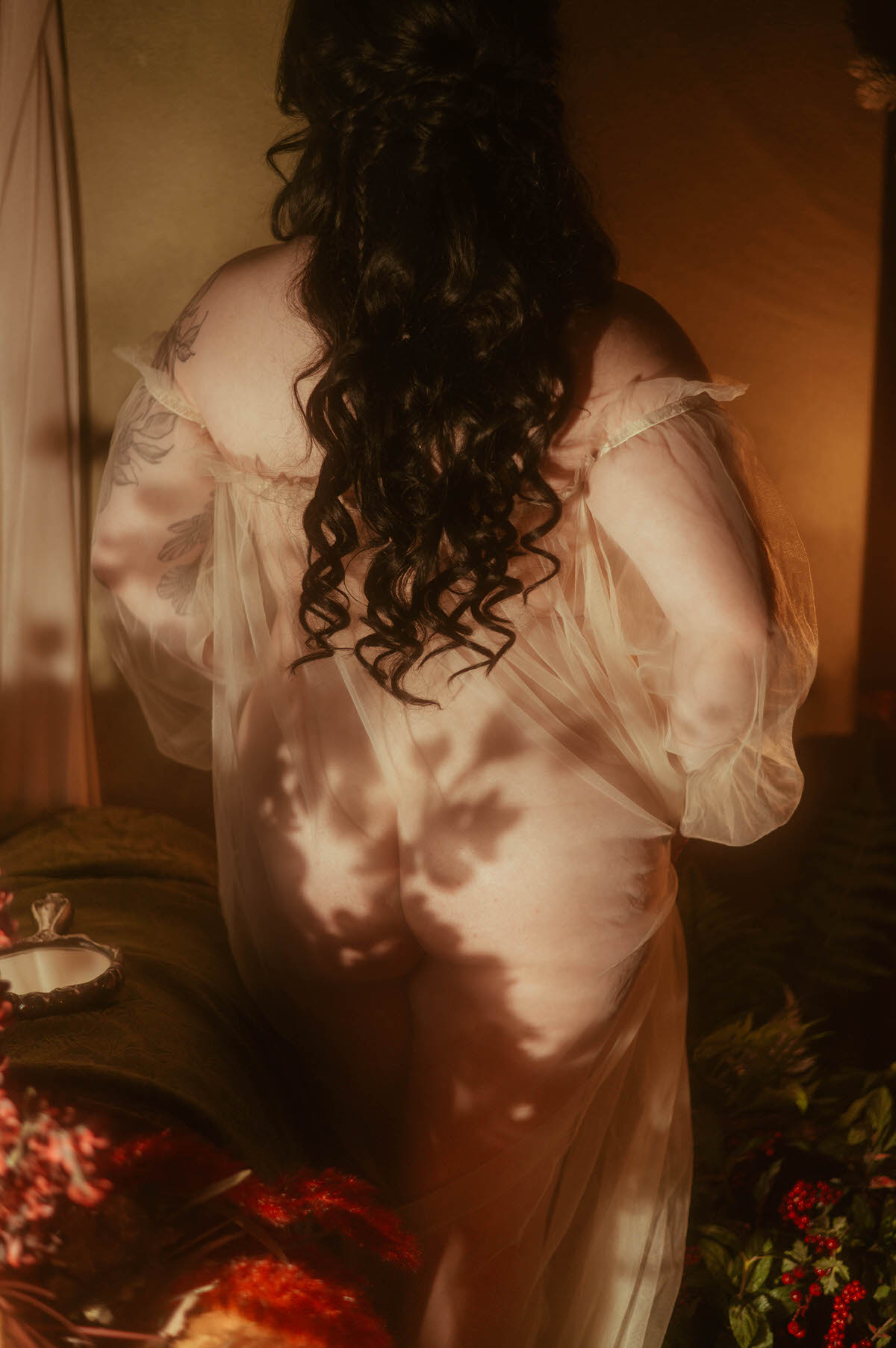 Back view of a woman in a sheer romantic gown, softly illuminated by golden light at Royal Lune Studio. Her long dark curls cascade down her back as she stands near a bed adorned with a vintage hand mirror, surrounded by fall florals and lush greenery. The dreamy boudoir scene evokes intimacy and softness through light, texture, and shadows.