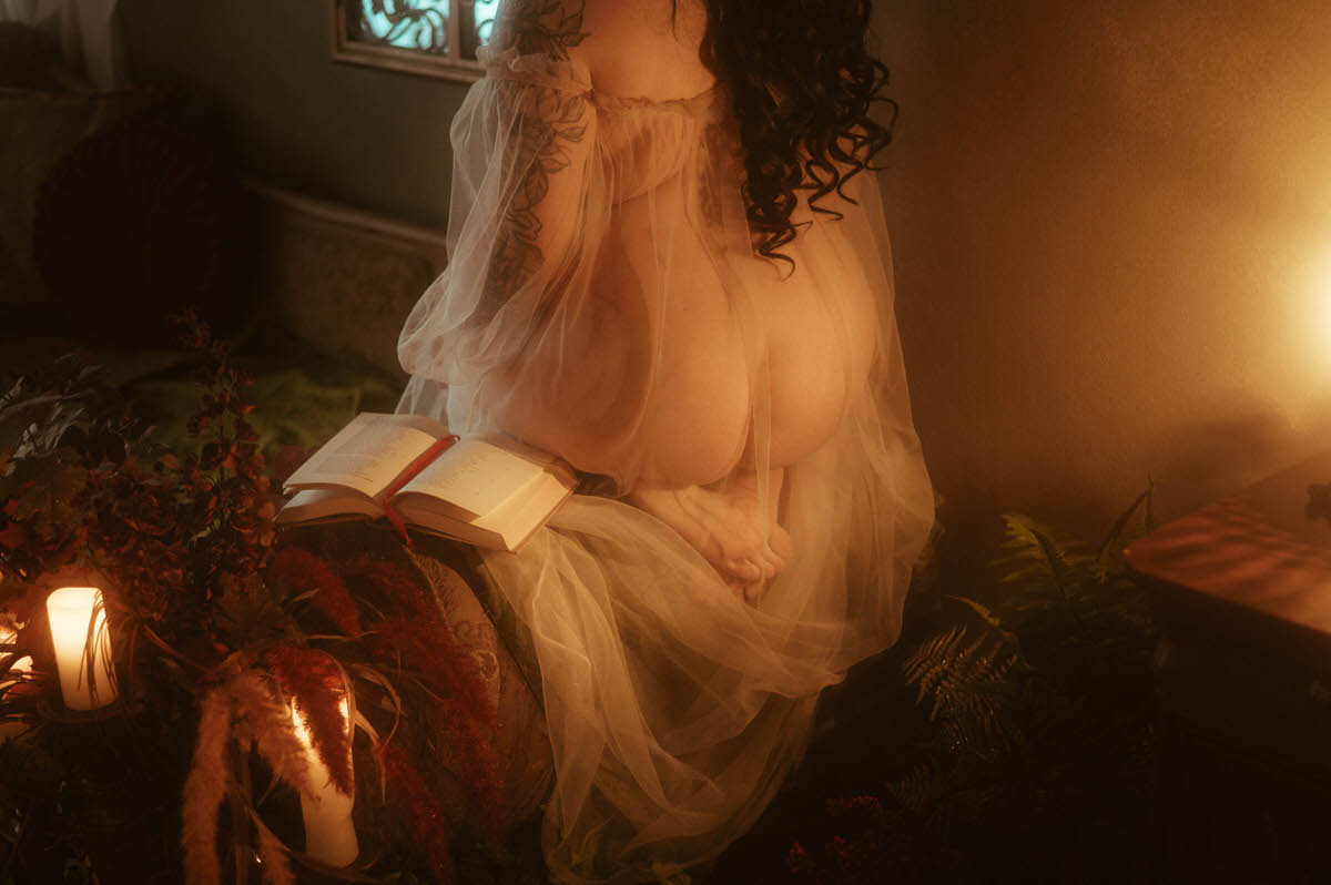 Close-up of a woman kneeling on a vintage couch in sheer fabric, her back turned to the camera with long dark curls and visible floral tattoos. Surrounded by candles, florals, and open books, the golden lighting adds intimacy and vintage charm to this moody boudoir photography session at Royal Lune Studio in Dallas.