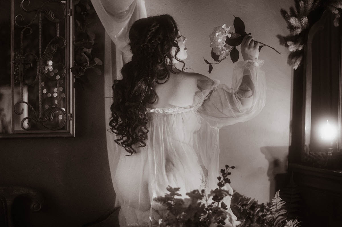 A woman in a sheer vintage gown stands with her back turned, holding a single flower near her face while soft candlelight glows beside her. Her long curls and romantic pose create a dreamy, painterly effect in this moody boudoir photography scene, styled with creative shadow play and vintage decor at a moody photography studio in Dallas.