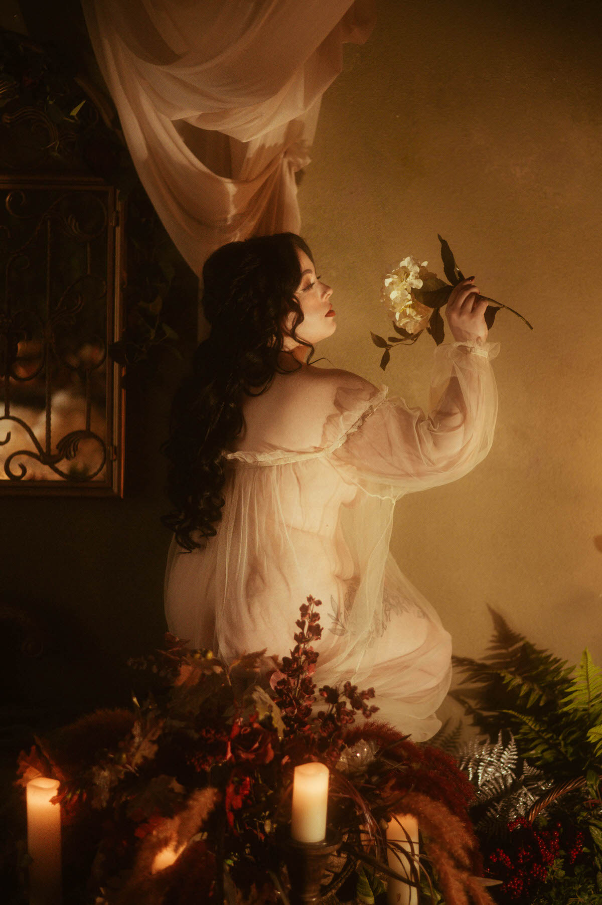 A curvy woman poses in profile wearing a sheer vintage robe, holding a white flower near her face while soft candlelight casts warm shadows across the wall. Surrounded by moody florals and antique decor, this dreamy boudoir photography scene captures romantic, creative inspiration at a moody photography studio in Dallas.