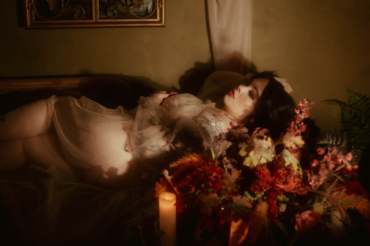 A woman lounges on a vintage couch in a sheer gown, illuminated by soft candlelight and surrounded by dried florals in rich autumn tones. Her pose and expression feel cinematic and vulnerable, capturing the essence of moody boudoir photography with artistic shadow play and vintage inspiration at a moody photography studio in Dallas.