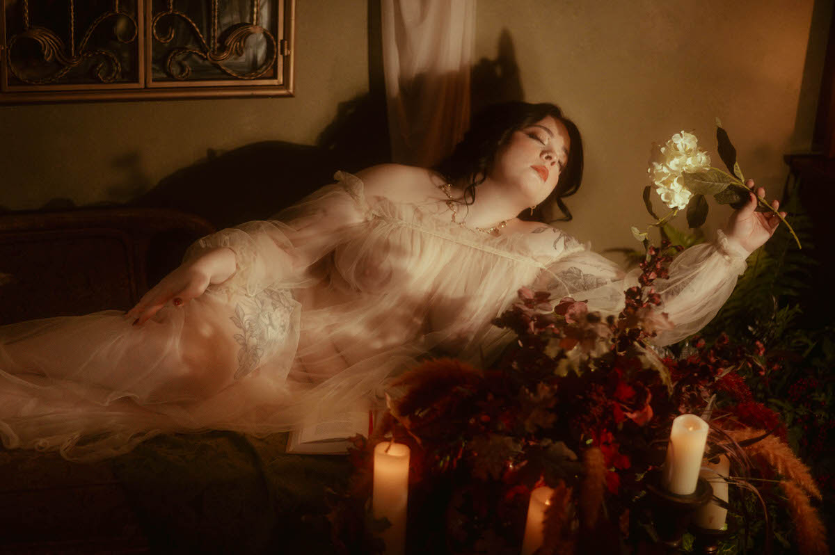 A woman reclines on a vintage couch at Royal Lune Studio, draped in a sheer romantic gown, holding a white flower in one hand with her eyes closed. Surrounded by autumn florals, open books, and glowing candlelight, the dreamy boudoir scene evokes Renaissance and Pre-Raphaelite influences through soft shadows and ethereal styling.