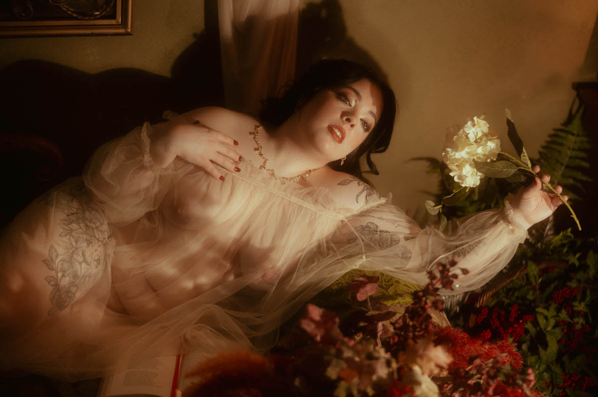 Reclining amidst dried florals and candlelight, a woman in a sheer gown gazes upward while holding a pale hydrangea stem. Her pose is soft and expressive, lit by golden shadows that create an intimate, painterly effect—an exquisite example of moody boudoir photography with vintage tones and creative boudoir inspiration at a moody photography studio in Dallas.