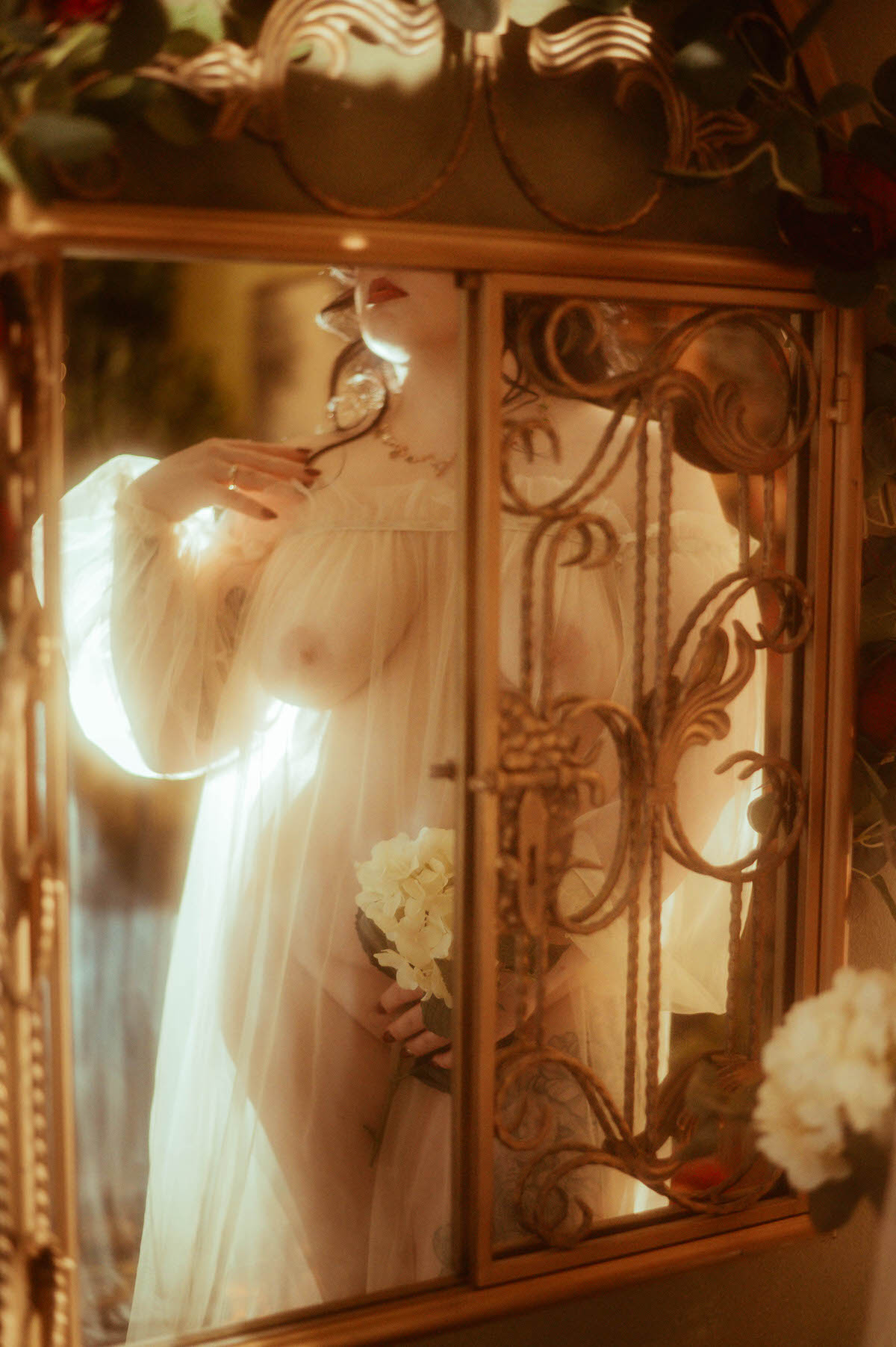 Artistic boudoir photo captured through a gilded mirror at Royal Lune Studio, showing a partially obscured nude woman in a sheer robe holding white flowers. Soft golden lighting enhances the dreamy, romantic mood, with floral details and vintage textures adding to the ethereal, moody boudoir photography aesthetic.