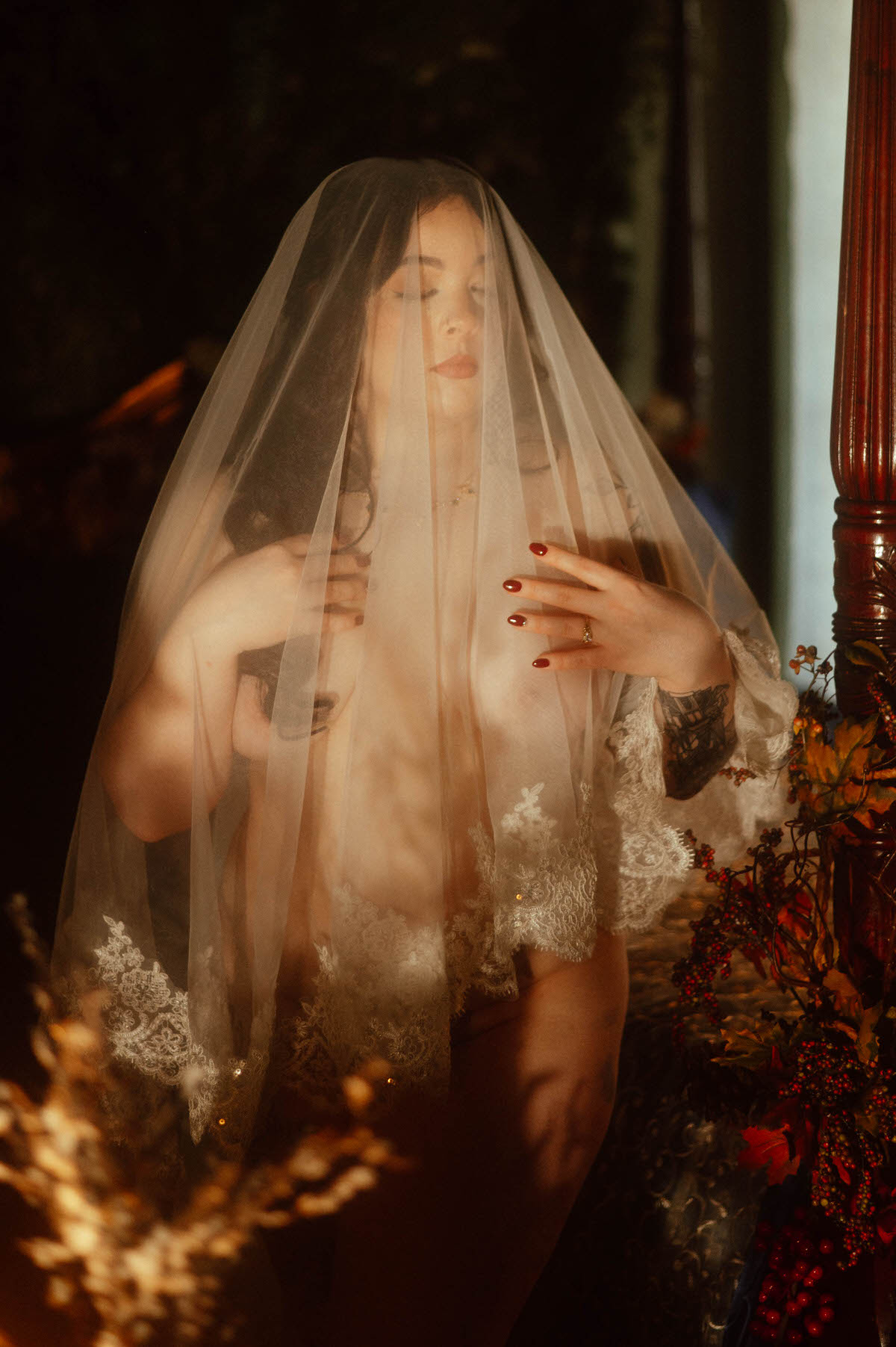 Artistic nude boudoir portrait of a woman draped in a sheer veil with lace trim, softly lit in golden tones at Royal Lune Studio. Her hands rest delicately over her chest, eyes closed, surrounded by warm fall foliage, evoking moody boudoir photography and vintage bridal fantasy.