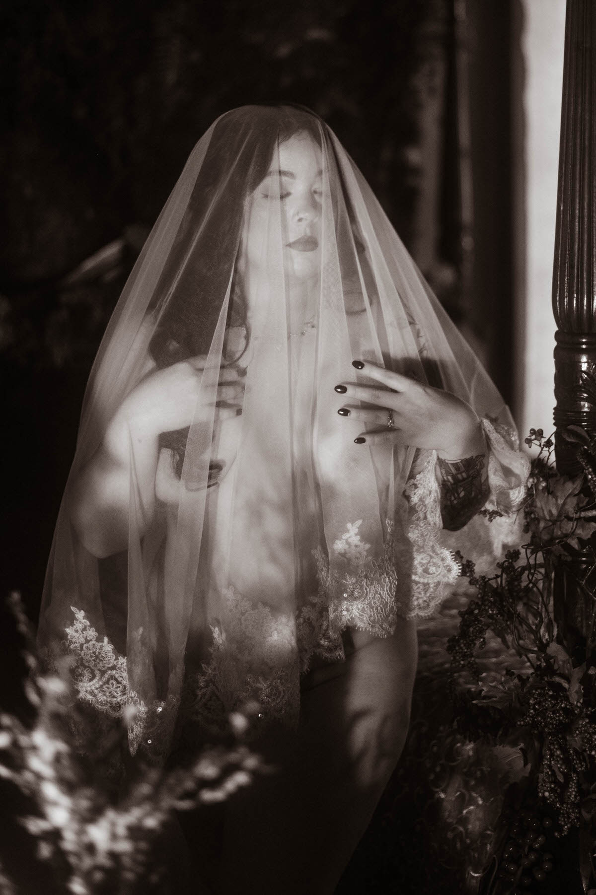 A nude woman stands beneath a sheer veil, holding a single white hydrangea, her tattoos softly visible through the fabric. The image’s moody boudoir photography style is enhanced by dramatic shadows and monochrome tones, evoking vintage romance and creative boudoir inspiration within a fine art setting.